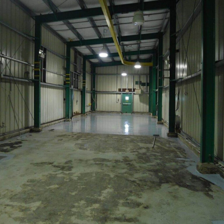 Walt Co Construction Interior view of an empty industrial warehouse with metal walls, concrete floors, and overhead lighting in Smooth Rock Falls, Ontario. A green door and some machinery can be seen in the background, suggesting recent work by Walt Co Construction. Smooth Rock Falls ON