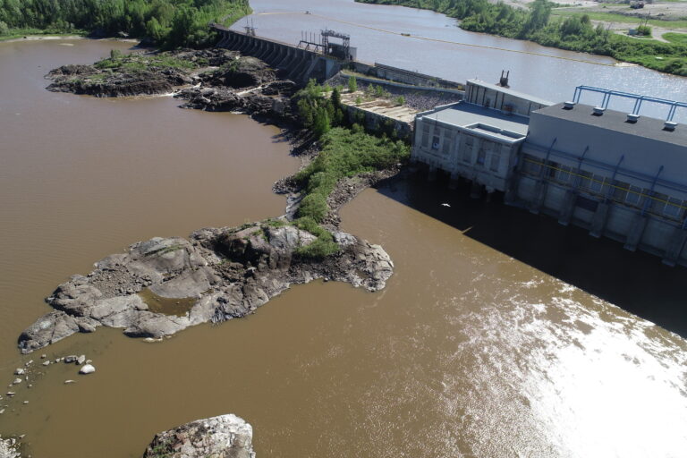 Walt Co Construction Aerial view of a dam with brown water flowing through it, surrounded by rocky terrain and green vegetation in Smooth Rock Falls, Ontario. The dam structure is visible on the right, with water on both sides, possibly hinting at the work done by Walt Co Construction. Smooth Rock Falls ON